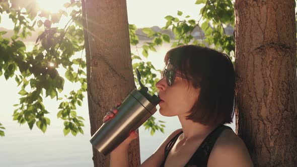 Sporty Woman Drinks From Metal Reusable Bottle Under Tree on the Riverbank alt