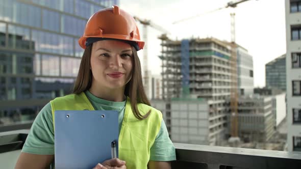 Portrait of a Young Female Builder in a Signal Vest and Helmet Against the Backdrop of a Building