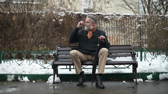 An adult gray-haired respectable man with glasses sits on a park bench alt