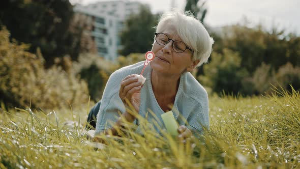Retired Senior Woman Enjoying Freedom. Childish Grandma Blowing Soap Bubbles in the Park alt