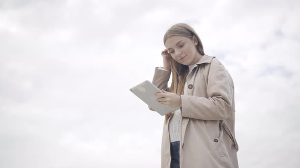 Bottom View of Happy Young Woman Using Tablet at the Background of Overcast Sky, Smiling and Hugging alt