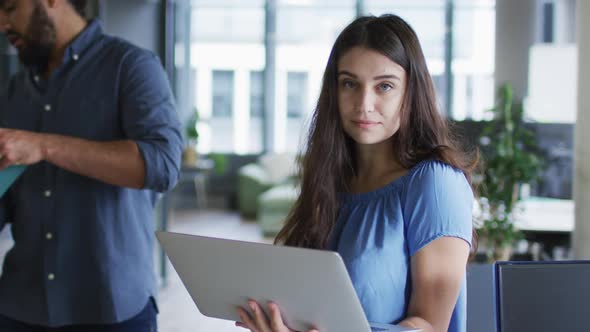 Portrait of caucasian businesswoman standing in office using laptop, looking to camera and smiling alt