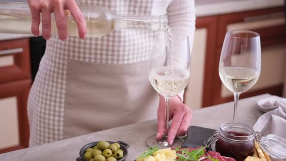 Woman Pouring Wine to a Glass From a Bottle at Domestic Kitchen with Meat and Cheese Plater on the alt