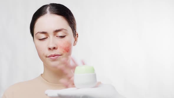 A young beautiful woman with a red cheek is offered a face cream, but she refuses. alt