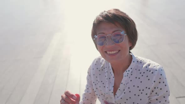 Woman with Colorful Sunglasses Sits on Scene in Urban Park and Enjoys of Bright Sunlight alt