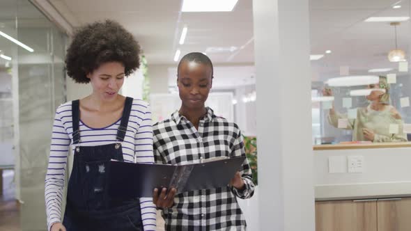Diverse smiling female business colleagues walking and discussing paperwork in office alt