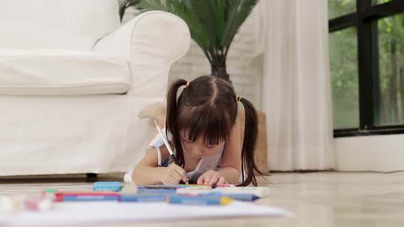 Cute little Asian girl lying on the floor drawing and painting on paper at home. alt