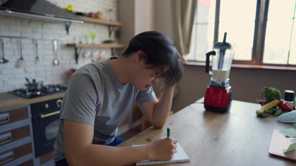Side View of Concentrated Millennial Man Writing Ideas in Sketchpad with Pen Sitting at Kitchen alt