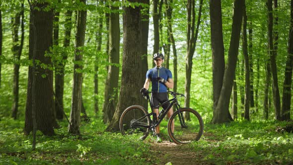 A Cyclist with a Smartphone in His Hands is Standing in the Middle of the Forest alt