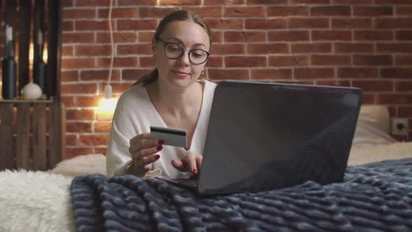 Caucasian Woman in Glasses is Lying on Bed and Uses Laptop and Bank Card to Pay for Purchases in alt