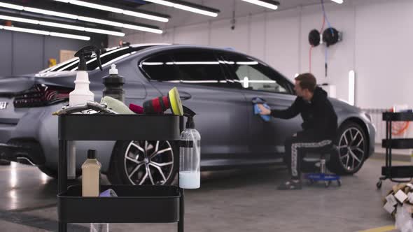 Auto Detailing Salon Man Worker Sitting Near Light Grey Car Polishing Surfaces Cleaning Devices alt