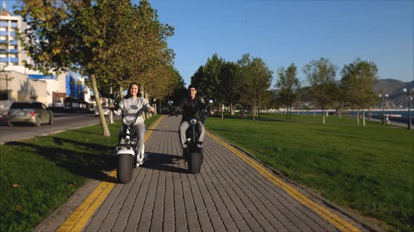 Man and Girl on Electric Scooters Are Riding Through Publick Park at Morning alt