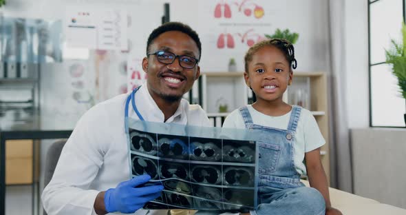 Doctor which Looking at Camera Together with Small Girl and Holding in His Hands Results of X-ray alt