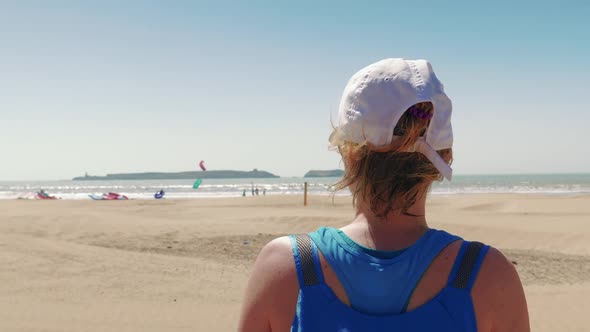 Tourist girl watching a beach full of kite surfers on sunny day, Morocco alt