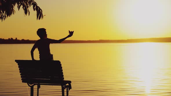 Little Boy is Sitting on the Bench and Playing with a Paper Boat  alt