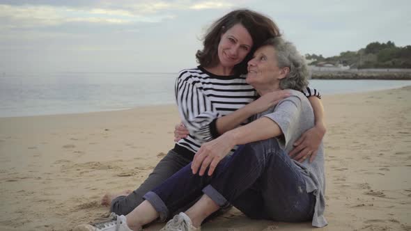 Laughing Daughter Hugging Old Mother While Sitting on Beach Together alt