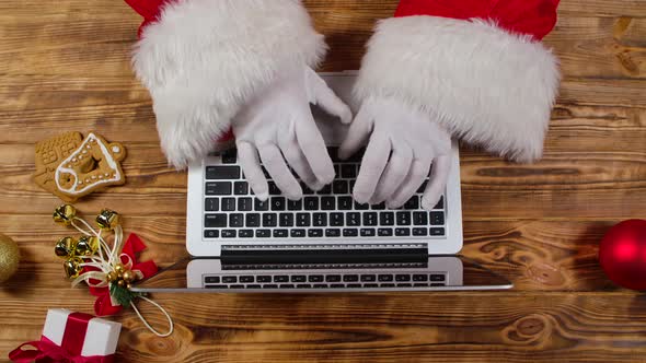 Top View Santa Hands in White Gloves Typing on the Keyboard By Wooden New Year Decorated Table alt