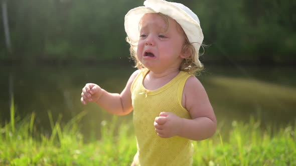 Little Funny Cute Blonde Girl Child Toddler in Yellow Bodysuit and White Hat Crying Walking in Field