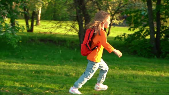 Little Preschooler Girl with School Backpack Runs and Laughs on Green Lawn in Summer Park