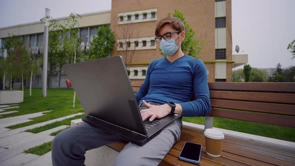 Yyoung man wearing glasses, siting on a bench outdoor and working on laptop computer. alt