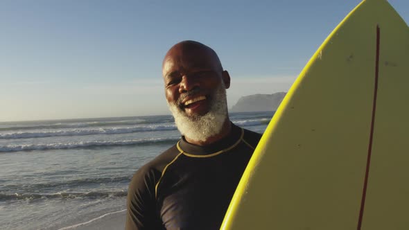 Smiling senior african american man walking with a surfboard at the beach alt