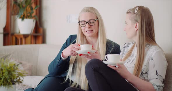 Two Women Drinking Coffee in Living Room Talking and Smiling. alt