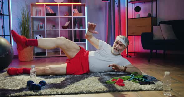 Old Bearded Man in Sportswear Doing Leg Stretching Exercises Lying on the Floor alt