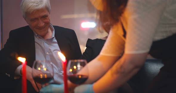 Waiter in Gloves Serving Red Wine at Table for Aged Man and Woman alt