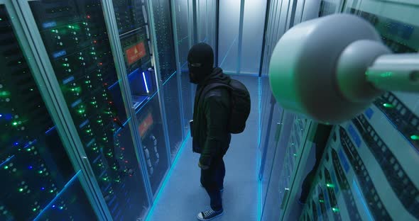 Masked Hacker with His Laptop Waking Through Server Rows alt