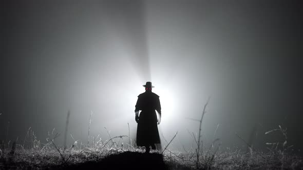 Silhouette of Plague Doctor Man in Long Mantle Costume and Hat Is Walking Ahead Through Smoke To alt