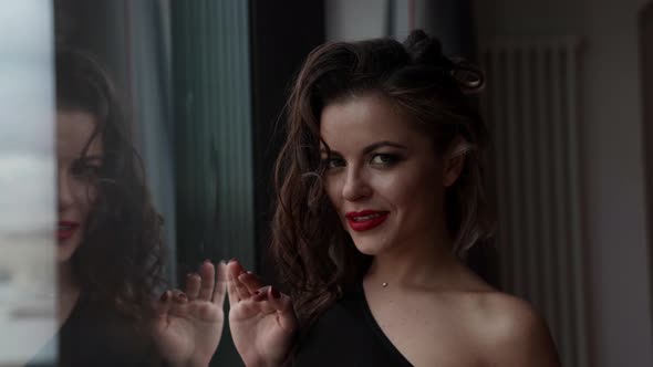 Portrait of Alluring Brunette Lady Near Window in Modern Apartment Flirting with Camera alt
