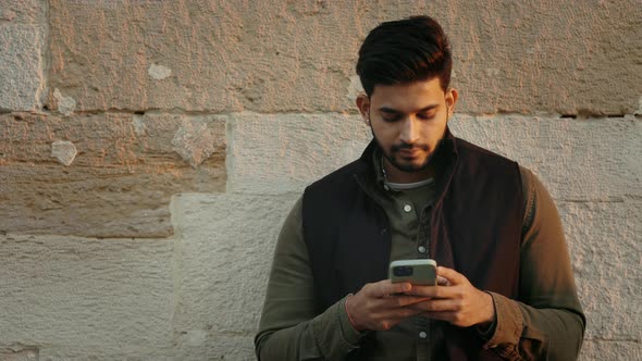 Indian Guy Using Smartphone While Standing Near Old Wall alt