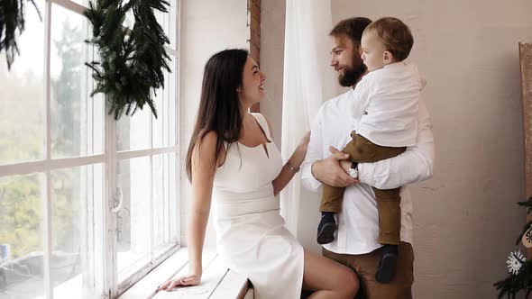 Young Father is Holding His Baby Boy Standing By the Window While Attractive Mother in White Dress alt