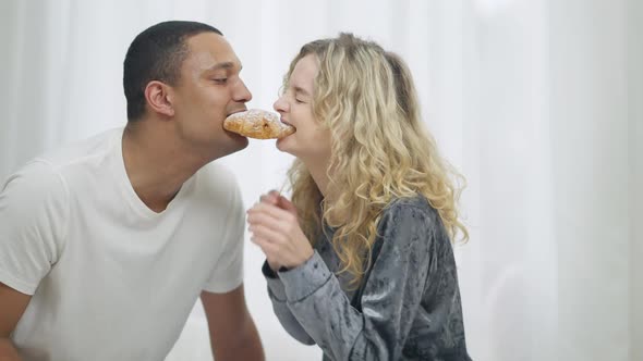 Side View of Cheerful Millennial Interracial Couple Eating Croissant From Both Sides in Bedroom in alt
