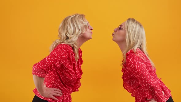 Two Blonde European Women Leaning Forward to Kiss Medium Shot Orange Background Studio Shot alt