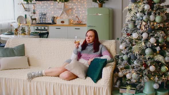 Woman Sitting on Sofa Near Christmas Tree and Drinking Alcoholic Beverage alt