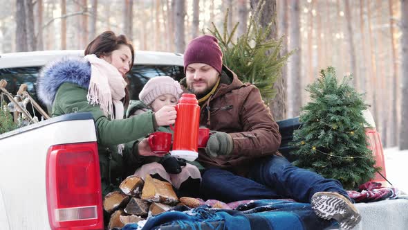 Happy Family of Three People Is Drinking Hot Tea From a Thermos, Sitting in the Back of a Car Near alt
