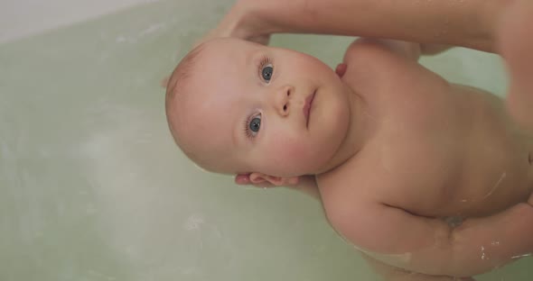 Calm Infant Lying in a Bath and Looking Up with Smile While Mother Washing Him alt