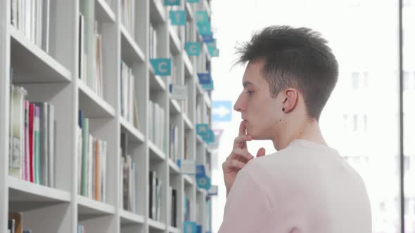 Young Man Looking Thoughtfully at the Bookshelves at the Library