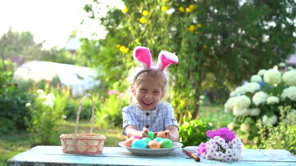a Little Blonde Girl with Bunny Ears Puts Colored Easter Eggs in a Basket in Nature and Smiles alt