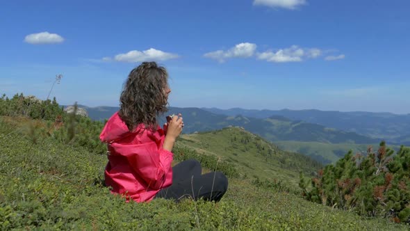 The girl on the top in the mountains is resting and drinking from a mug. 