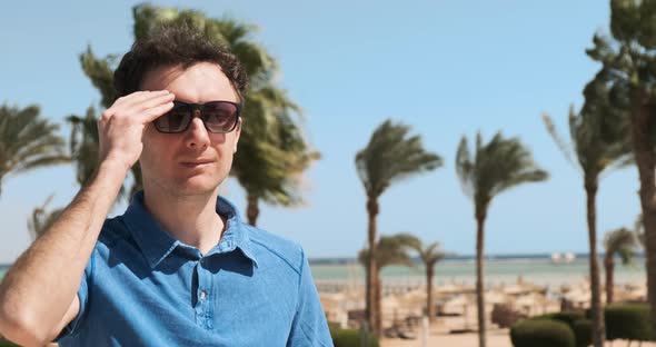 Man in Blue Shirt on Sun Vacation Wears Sunglasses Drinks an Orange Cocktail Backdrop of Palm Trees alt