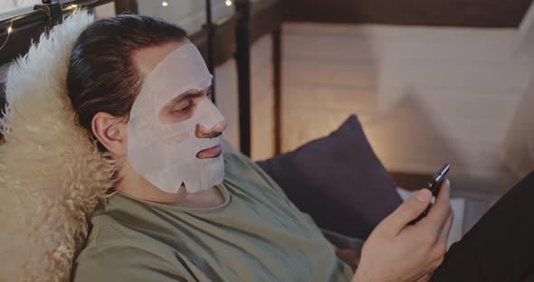 A Man in a Tissue Moisturizing Mask is Resting on a Bed or Sofa with a Phone