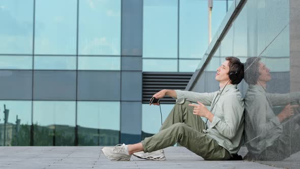 Happy Millennial Guy in Earphones Sitting on Ground in Front of Modern Glass Building Enjoy Rhythmic alt