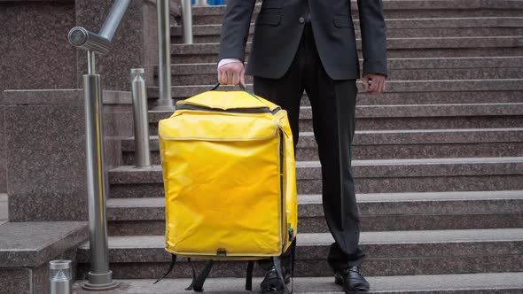 Unrecognizable Man in Formal Suit Standing with Yellow Thermal Backpack on Urban Stairs Outdoors alt