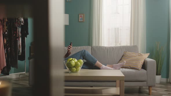Young Woman Eats a Red Apple Lying on Sofa and and Watching Something Gadget