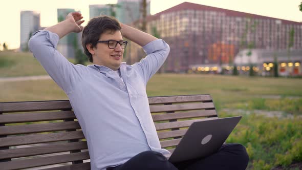 Young Man Finishes Work in Laptop Sitting on a Bench on Blurred Park Background