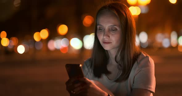 At Night, a Young Girl Holds a Smartphone in Her Hands and Looks at the Screen alt