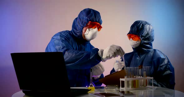 People in Protective Clothing with Hoods, Respirators, Glasses and Gloves Sit at a Table with a alt
