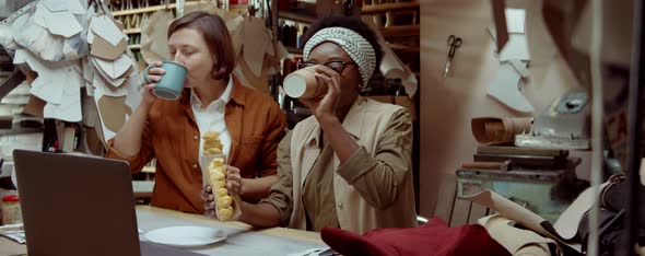 Women Drinking Coffee, Eating Pastry and Using Laptop in Workshop alt